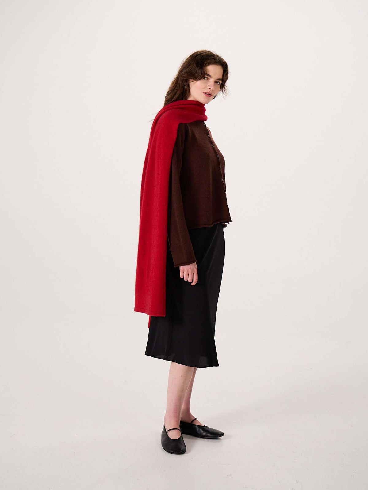 A woman stands in profile against a plain light background, wearing a brown top, black skirt, black shoes, and the Umbra Scarf – Poppy—a long red scarf measuring 22cm wide by 97cm long—draped over her shoulders.