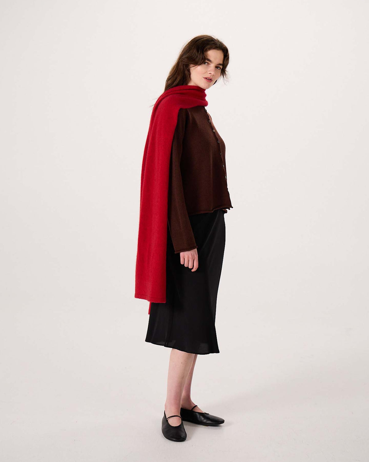 A woman stands in profile against a plain light background, wearing a brown top, black skirt, black shoes, and the Umbra Scarf – Poppy—a long red scarf measuring 22cm wide by 97cm long—draped over her shoulders.