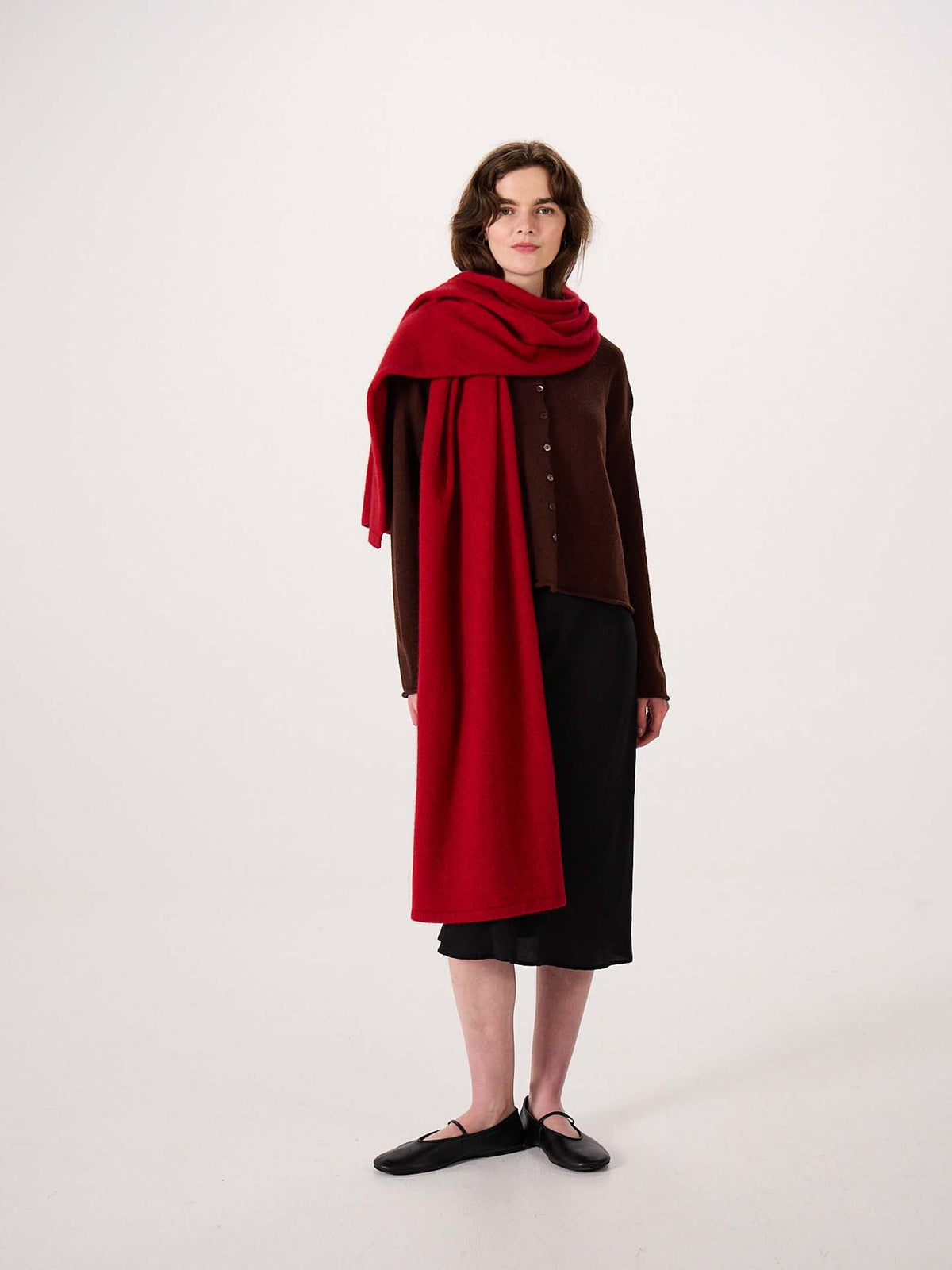 A person stands against a plain white background, wearing a brown buttoned cardigan, black skirt, black shoes, and the Umbra Scarf – Poppy—a large red scarf measuring 22cm wide and 97cm long—draped over one shoulder.