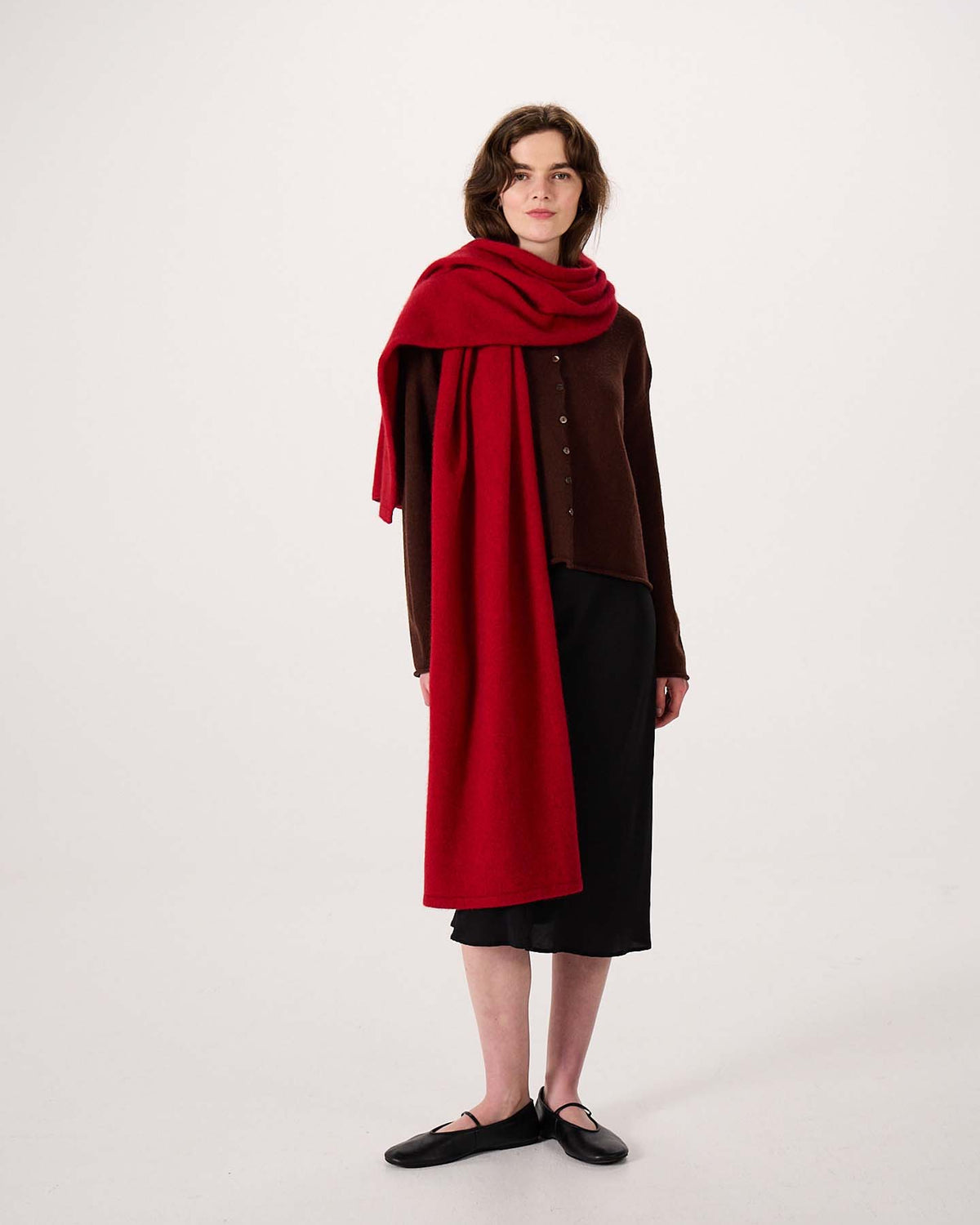 A person stands against a plain white background, wearing a brown buttoned cardigan, black skirt, black shoes, and the Umbra Scarf – Poppy—a large red scarf measuring 22cm wide and 97cm long—draped over one shoulder.