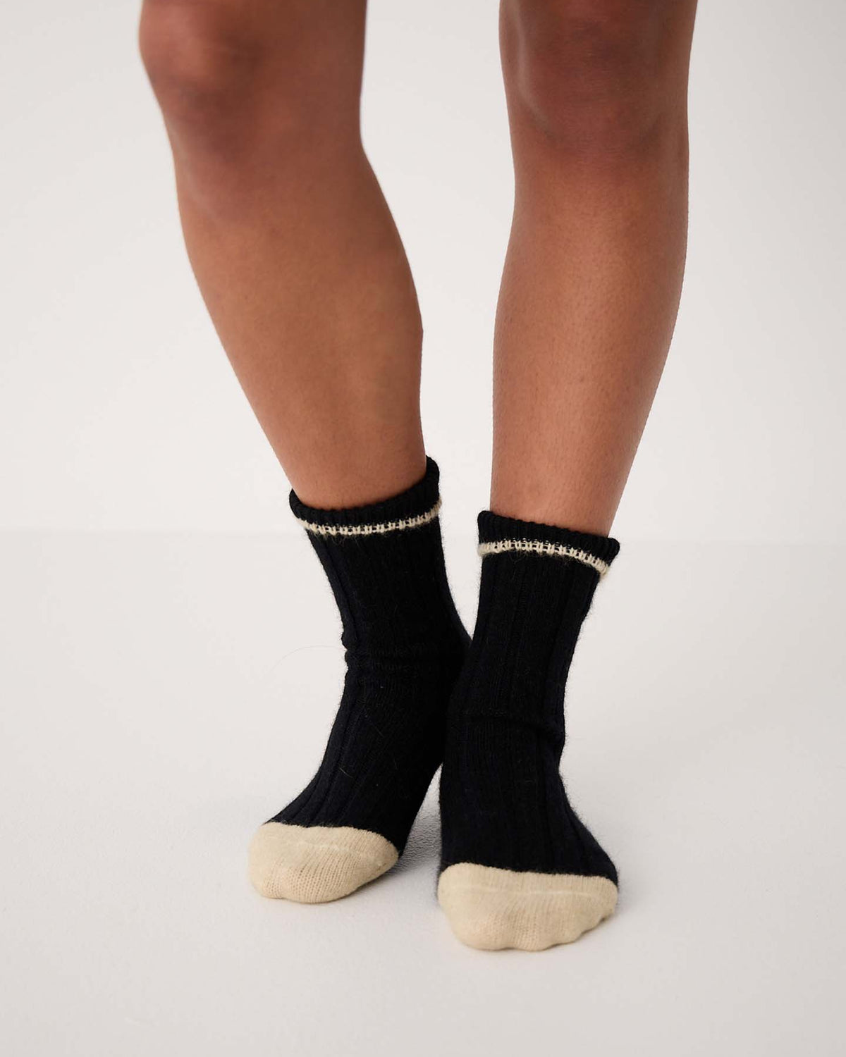 Someone is wearing Yin & Yang Socks – Black & Creme Toe (medium) with black ribbed fabric and beige toes and heels, standing on a white surface.