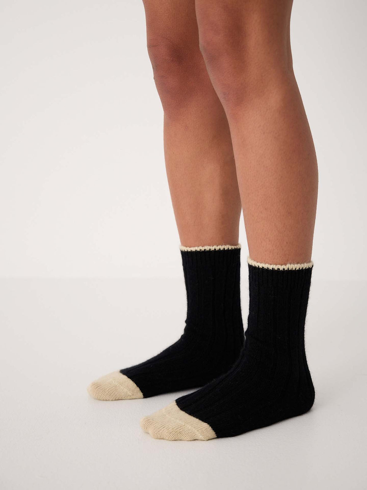 A person wearing Yin & Yang Socks – Black & Creme Toe, available in small, medium, and large, stands on a white floor against a plain white background.