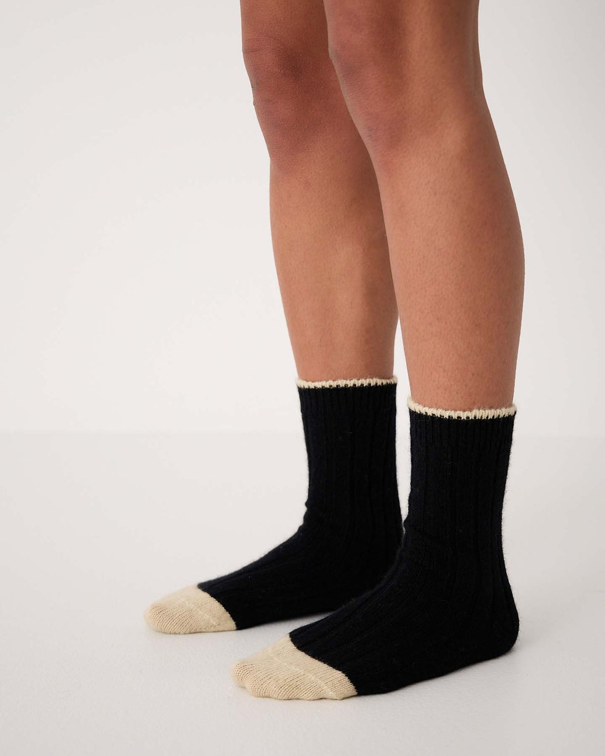 A person wearing Yin & Yang Socks – Black & Creme Toe, available in small, medium, and large, stands on a white floor against a plain white background.