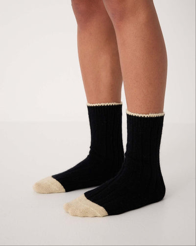 A person wearing Yin & Yang Socks – Black & Creme Toe, available in small, medium, and large, stands on a white floor against a plain white background.