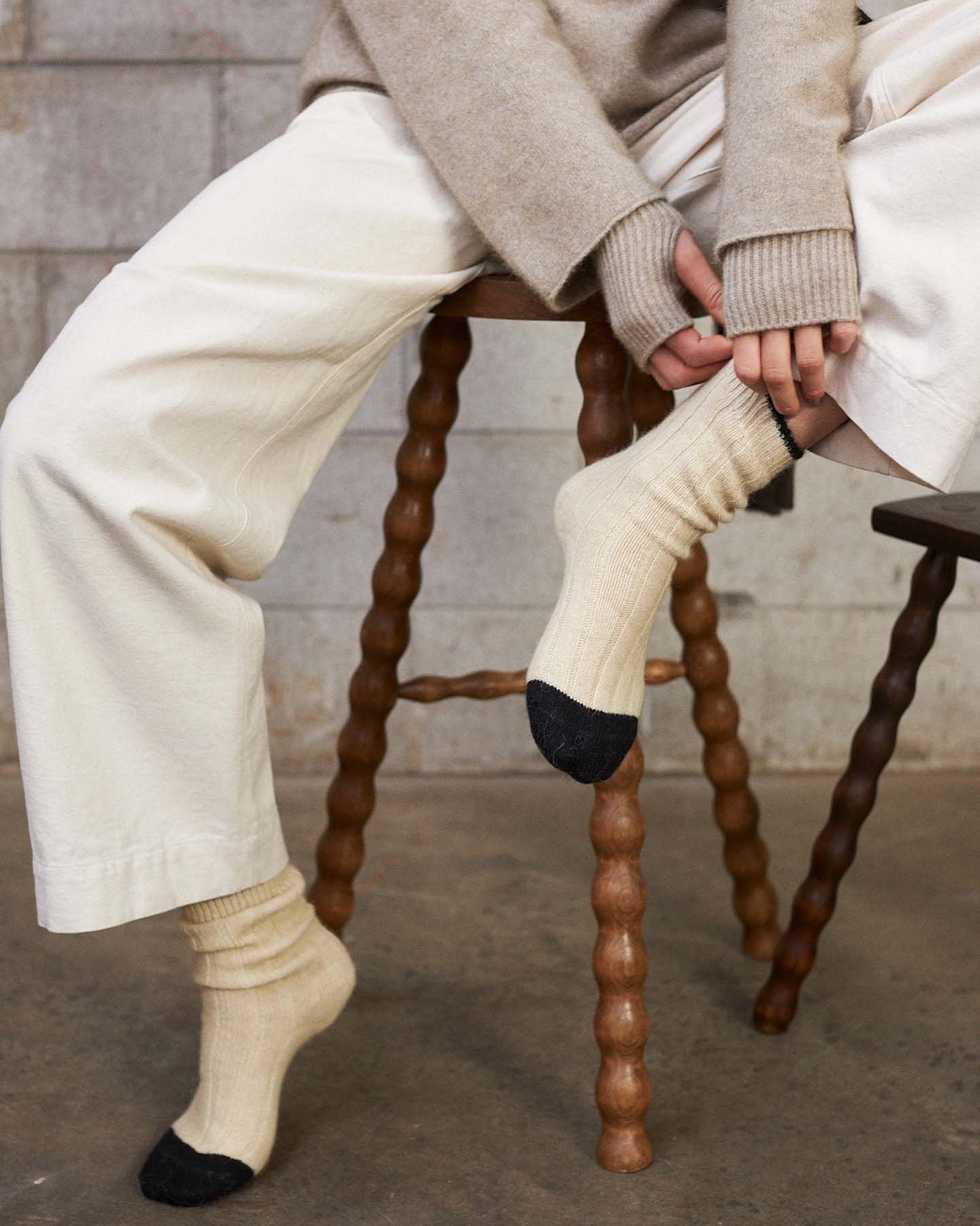 A person in cream pants and a sweater sits on a wooden stool, pulling up Yin & Yang Socks – Creme & Black Toe. The concrete wall and floor background highlights all sizes—small, medium, or large—per the shoe size chart.
