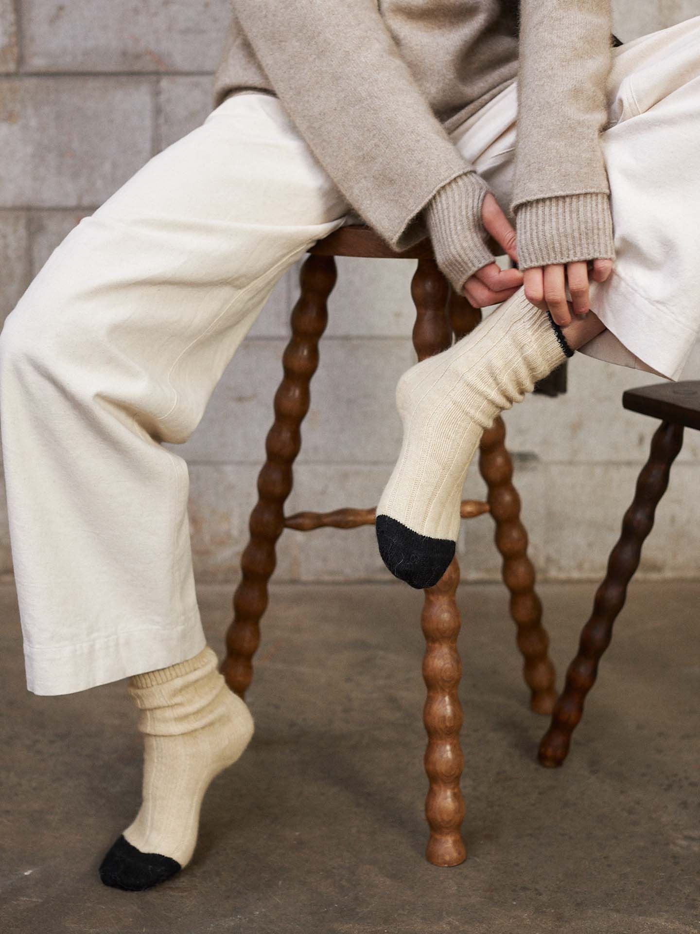 A person in cream pants and a sweater sits on a wooden stool, pulling up Yin & Yang Socks – Creme & Black Toe. The concrete wall and floor background highlights all sizes—small, medium, or large—per the shoe size chart.