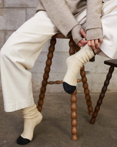 A person in cream pants and a sweater sits on a wooden stool, pulling up Yin & Yang Socks – Creme & Black Toe. The concrete wall and floor background highlights all sizes—small, medium, or large—per the shoe size chart.