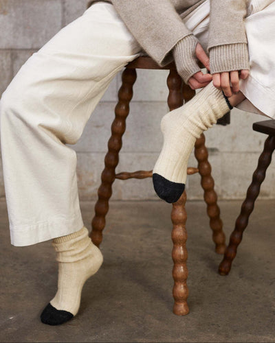A person in cream pants and a sweater sits on a wooden stool, pulling up Yin & Yang Socks – Creme & Black Toe. The concrete wall and floor background highlights all sizes—small, medium, or large—per the shoe size chart.