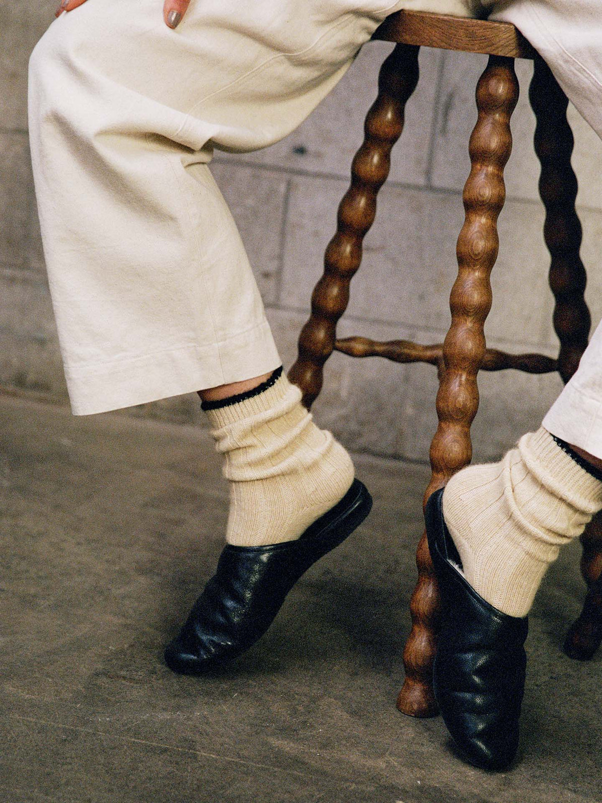 A person wears Yin & Yang Socks – Creme & Black Toe with cream pants and black shoes, seated on a twisted-leg wooden stool against a concrete wall—ideal for selecting sizes S, M, L or viewing the size chart (EUR, UK, US).
