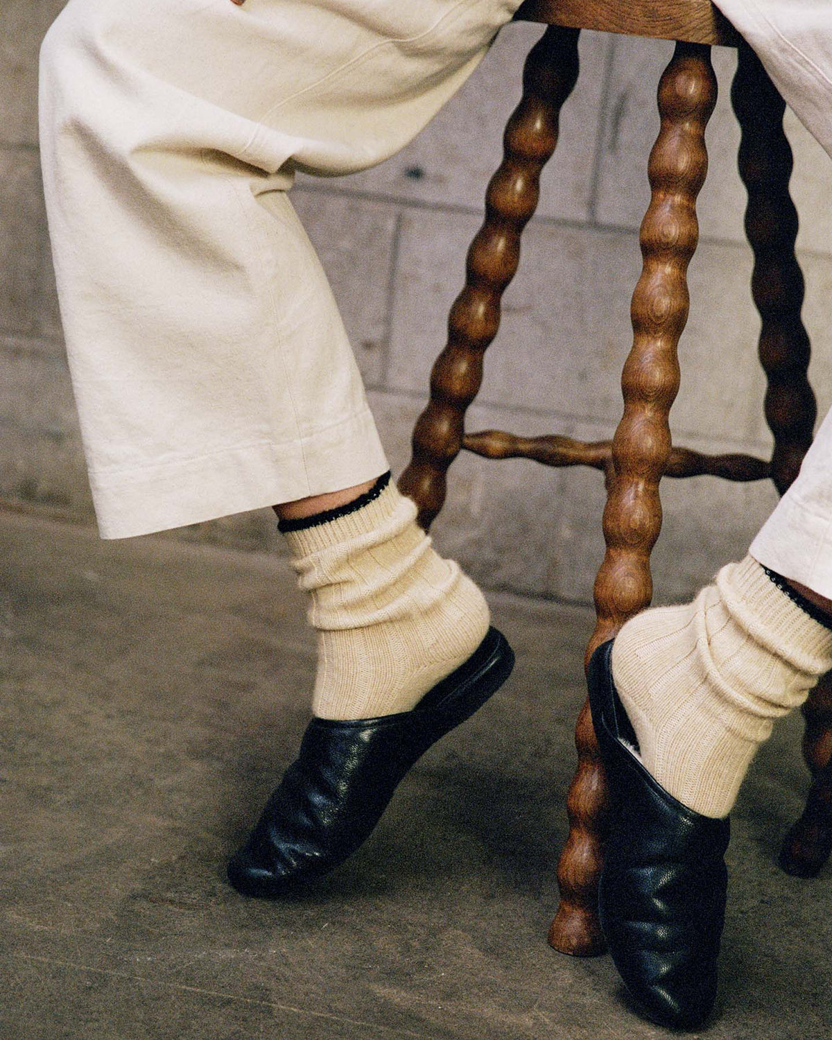 A person wears Yin & Yang Socks – Creme & Black Toe with cream pants and black shoes, seated on a twisted-leg wooden stool against a concrete wall—ideal for selecting sizes S, M, L or viewing the size chart (EUR, UK, US).