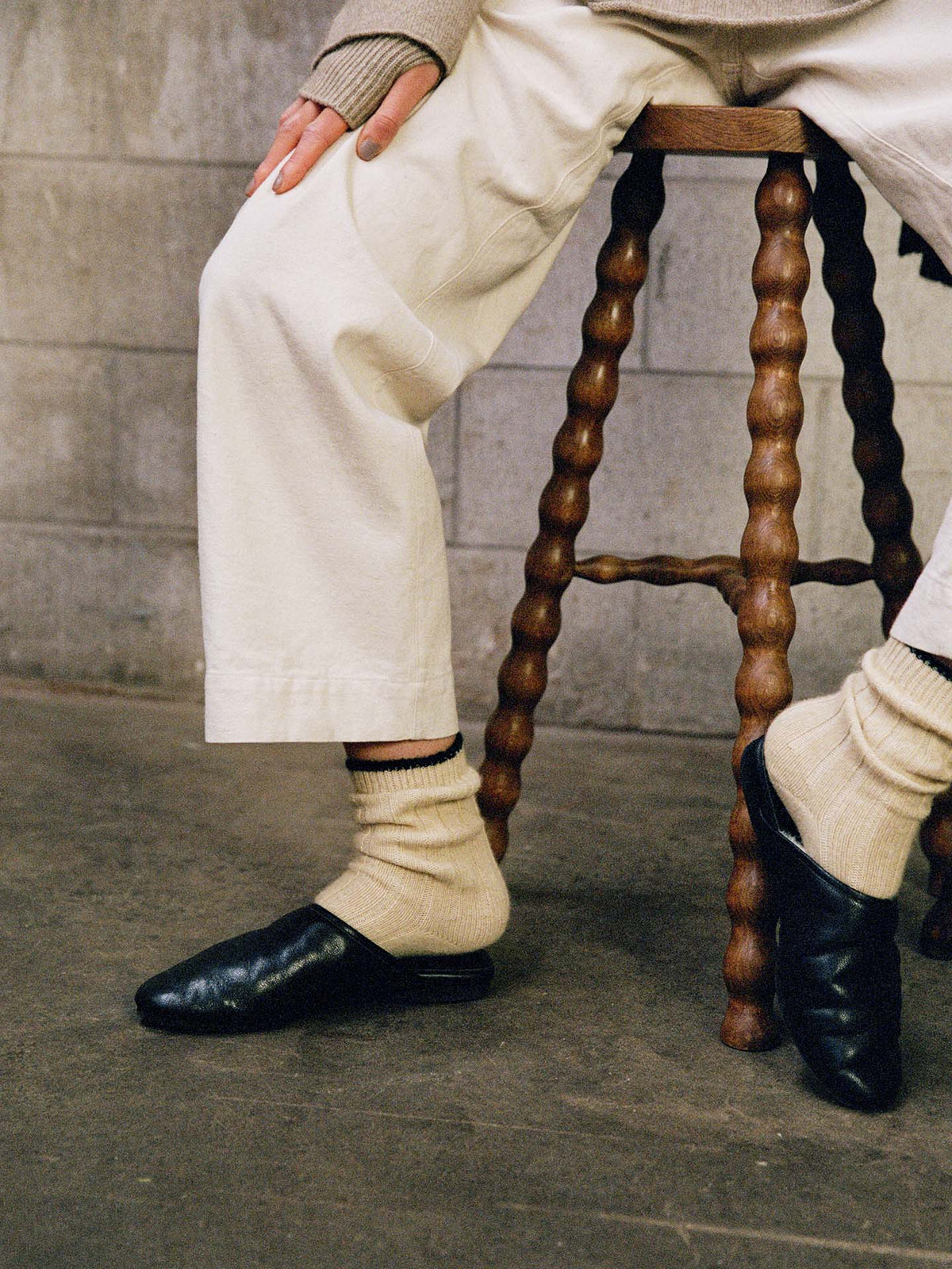 A person wearing white pants and Yin & Yang Socks – Creme & Black Toe with black slip-ons sits on a wooden stool against a concrete wall, highlighting the style’s versatile fit in small, medium, or large sizes via our shoe size chart.