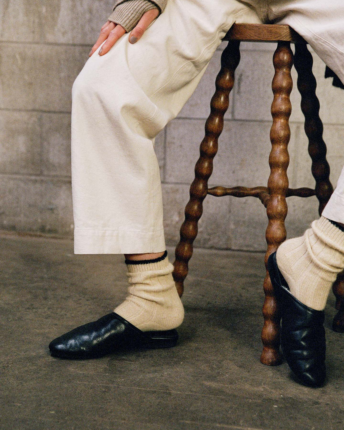 A person wearing white pants and Yin & Yang Socks – Creme & Black Toe with black slip-ons sits on a wooden stool against a concrete wall, highlighting the style’s versatile fit in small, medium, or large sizes via our shoe size chart.