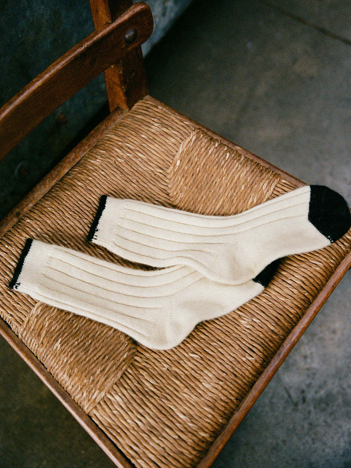The Yin & Yang Socks – Creme & Black Toe, featuring a cream ribbed design with black heels and toes, are displayed on a woven wooden chair and come in small, medium, and large per our shoe size chart.
