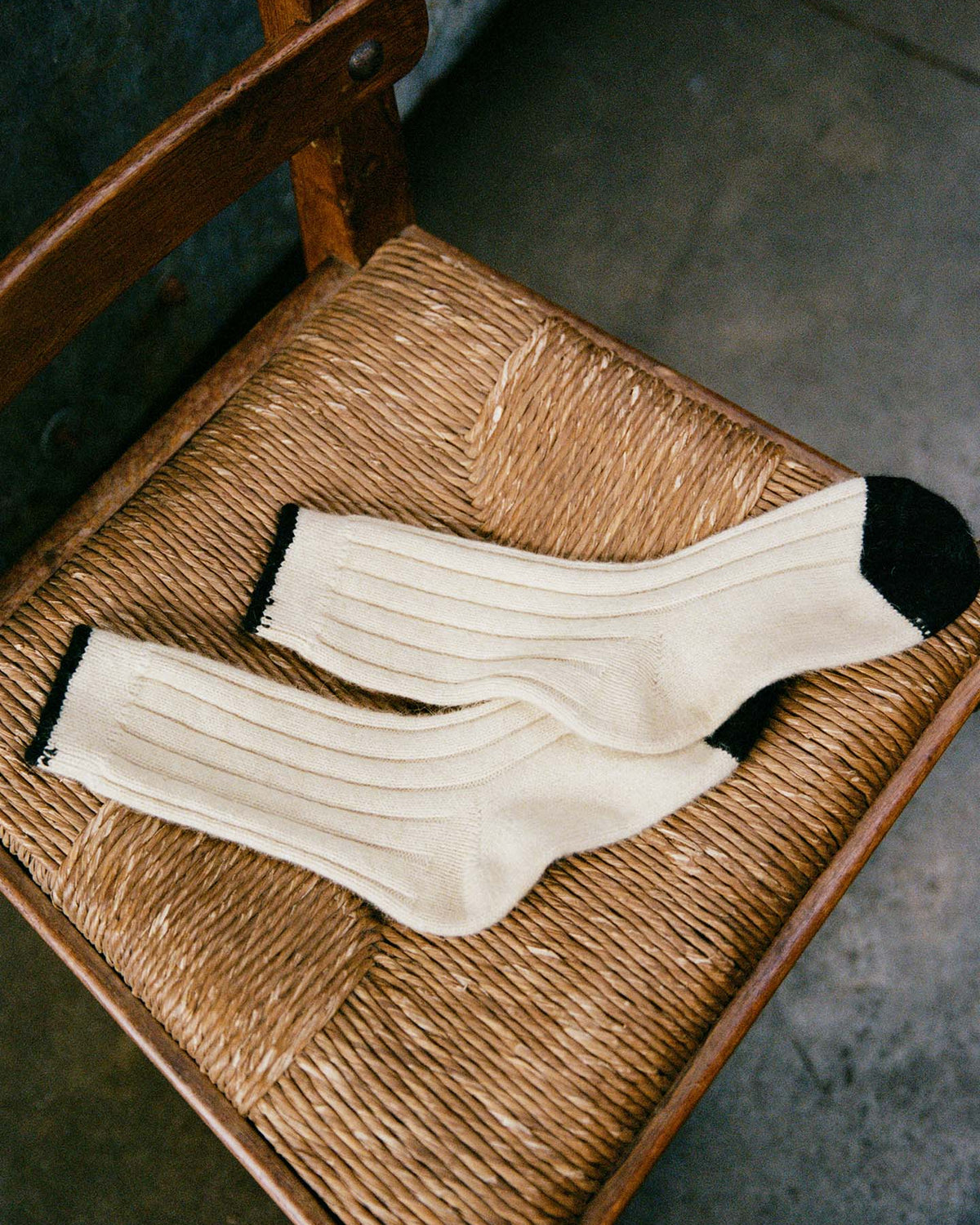 The Yin & Yang Socks – Creme & Black Toe, featuring a cream ribbed design with black heels and toes, are displayed on a woven wooden chair and come in small, medium, and large per our shoe size chart.