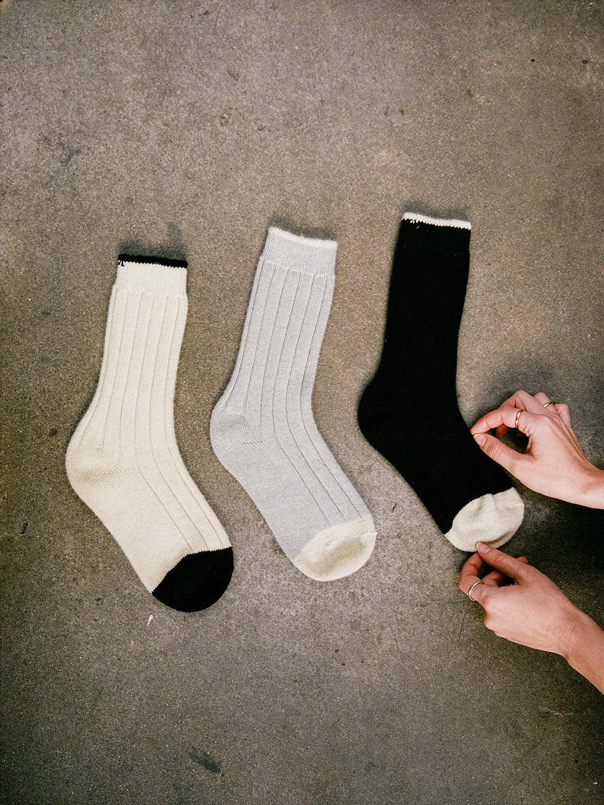 Three Yin & Yang Socks – Black & Creme Toe, in small, medium, and large—cream with black toe and heel, light gray, and black—are laid out on a concrete floor; a person adjusts the black sock.