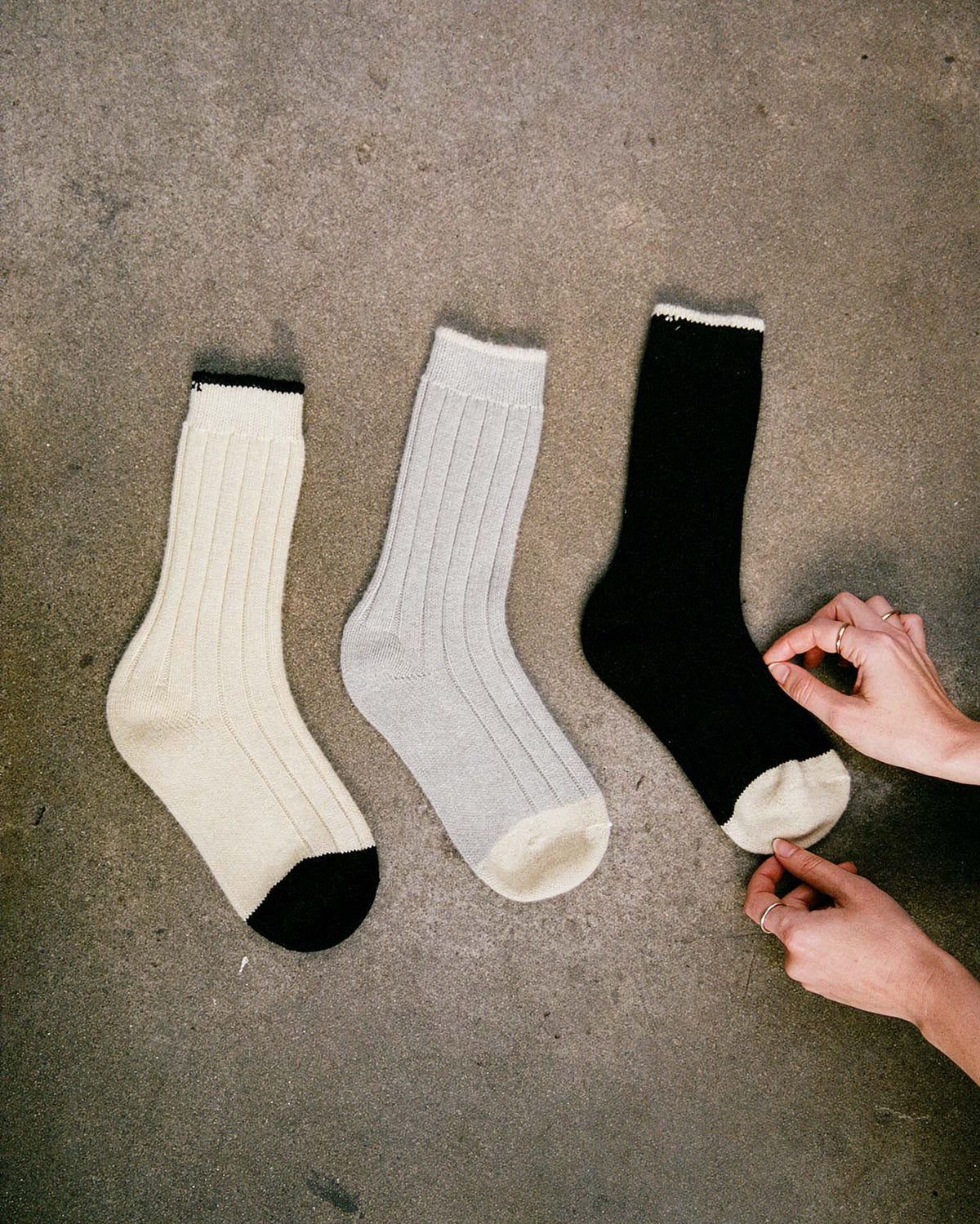 Three Yin & Yang Socks – Black & Creme Toe, in small, medium, and large—cream with black toe and heel, light gray, and black—are laid out on a concrete floor; a person adjusts the black sock.