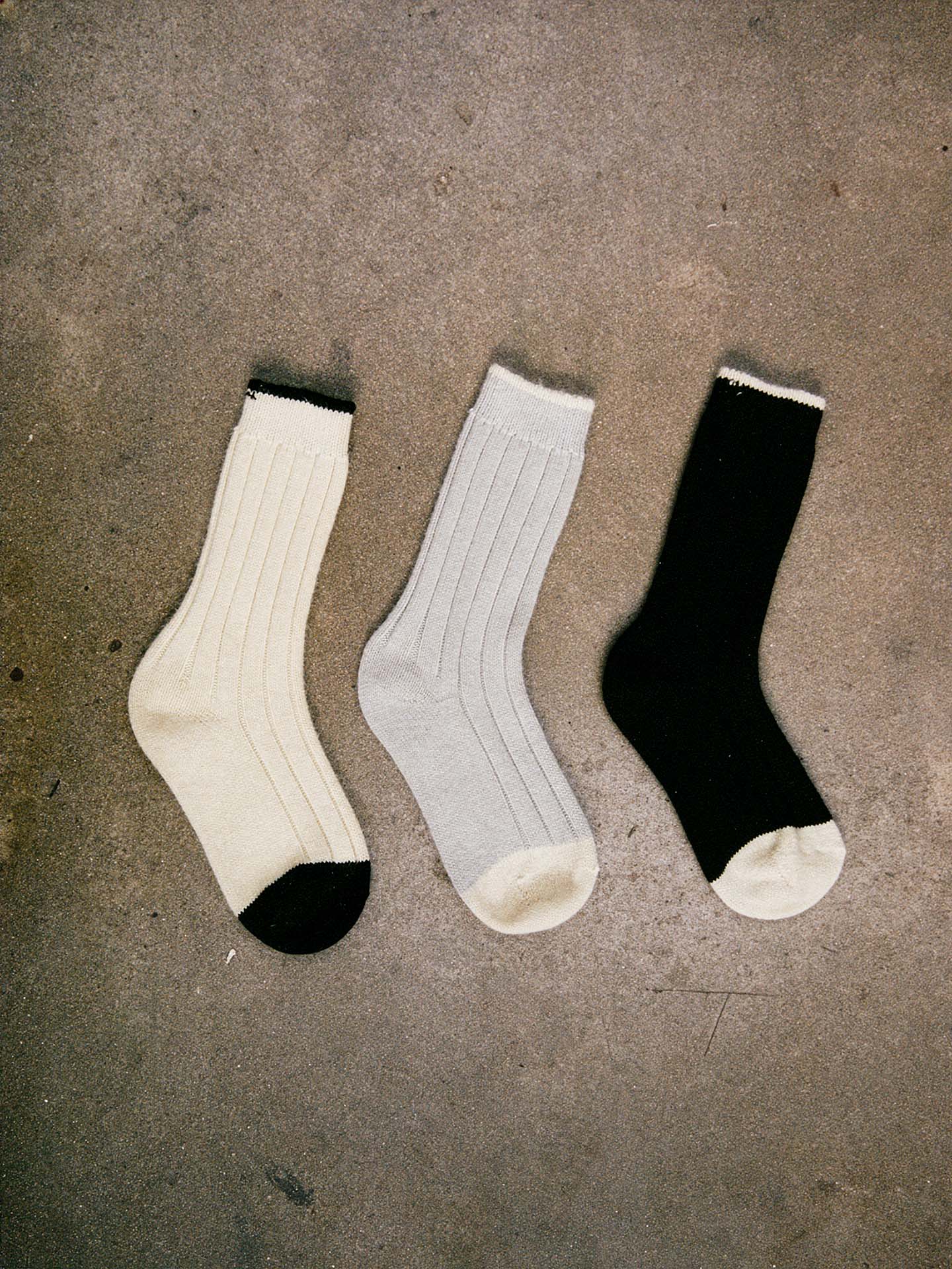 Yin & Yang Socks – Black & Creme Toe: Three ribbed socks in cream, light gray, and black with contrasting accents, displayed on textured concrete. Available in S, M, or L; see our shoe size chart for EUR, UK, and US sizing.