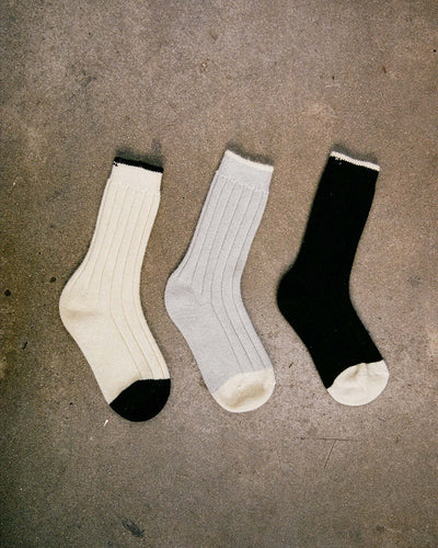 Yin & Yang Socks – Black & Creme Toe: Three ribbed socks in cream, light gray, and black with contrasting accents, displayed on textured concrete. Available in S, M, or L; see our shoe size chart for EUR, UK, and US sizing.