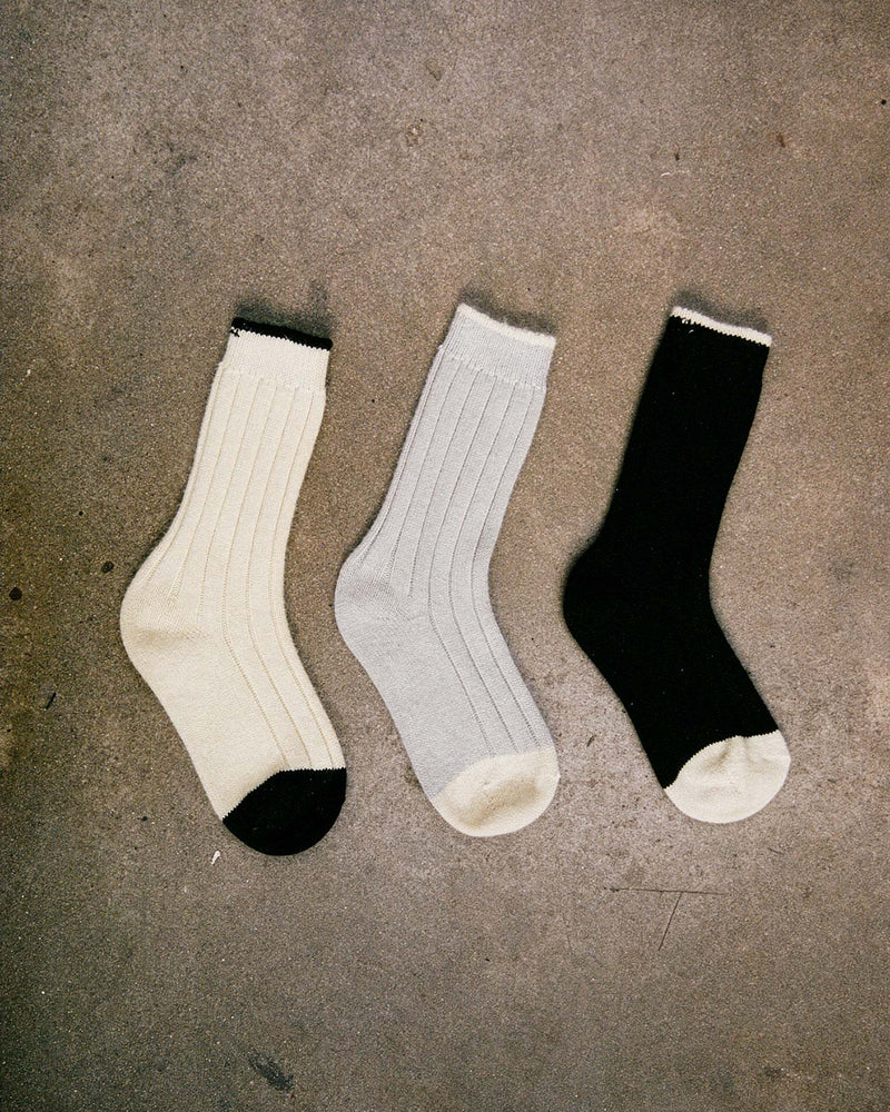 Yin & Yang Socks – Black & Creme Toe: Three ribbed socks in cream, light gray, and black with contrasting accents, displayed on textured concrete. Available in S, M, or L; see our shoe size chart for EUR, UK, and US sizing.