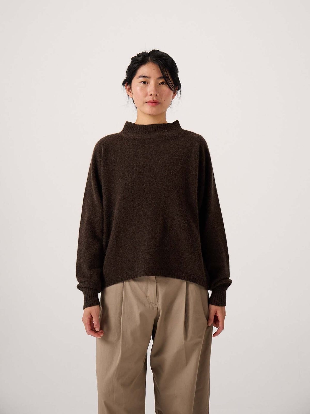 A person stands facing the camera against a plain light background, wearing the Fireside Knit – Truffle sweater in a relaxed fit with beige pants.