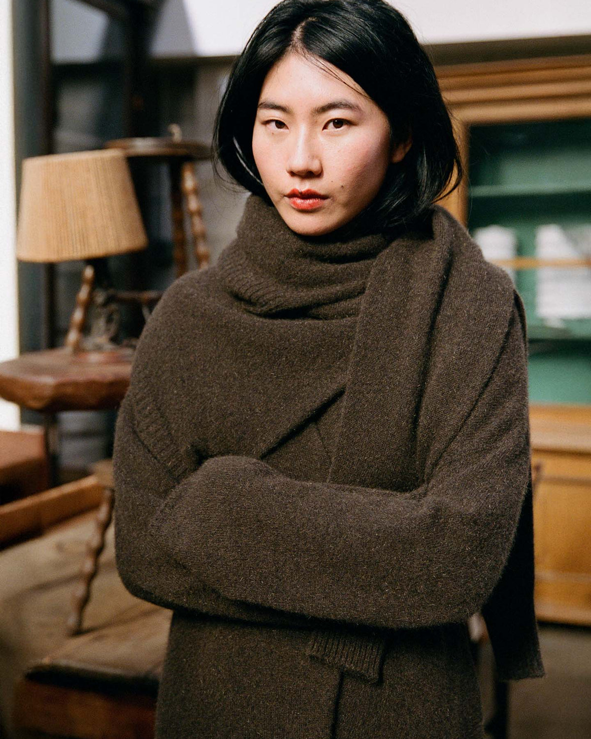 A woman with straight black hair stands indoors, arms crossed, wearing the Cocoon Coat – Truffle as she looks at the camera.