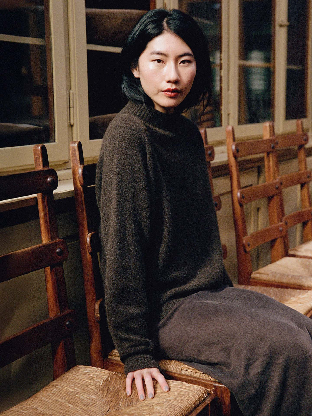 A person with straight black hair, wearing the Fireside Knit – Truffle in a relaxed fit and a skirt, sits on a wooden chair indoors next to empty chairs.