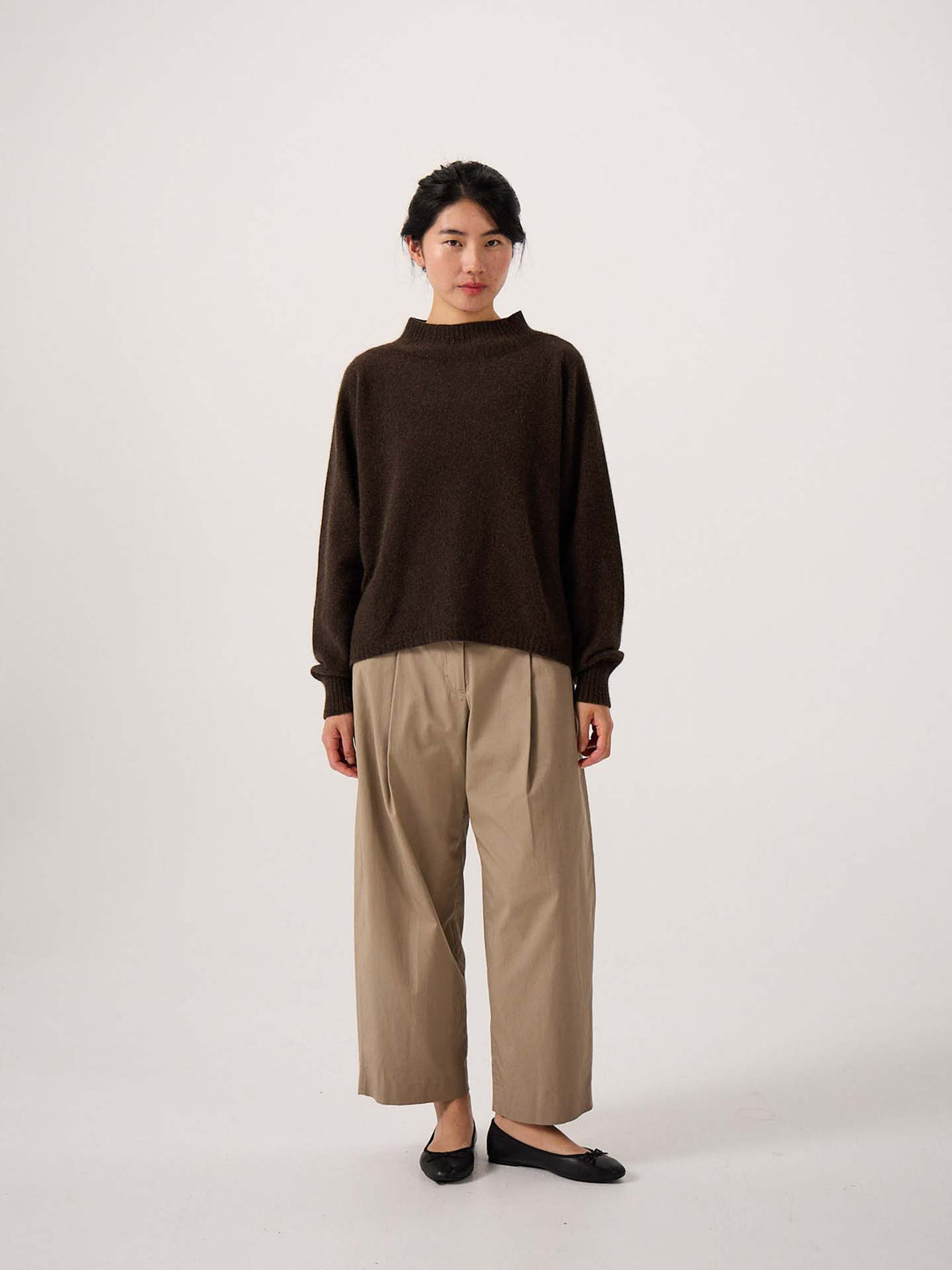 A woman stands against a plain white background wearing the Fireside Knit – Truffle, an oversized brown sweater, paired with beige wide-leg pants and black flats.