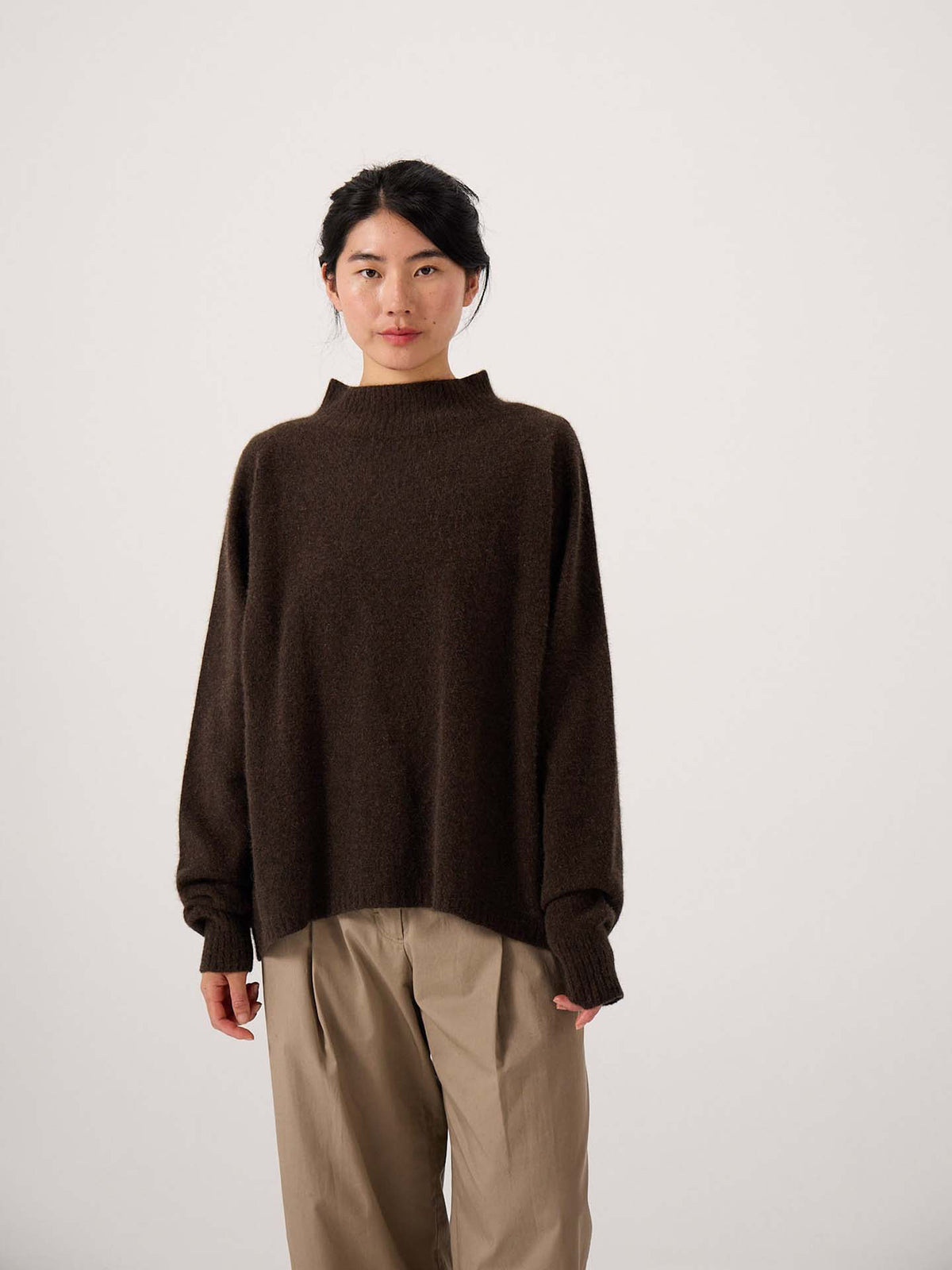 A person stands facing forward against a plain light background, wearing the Fireside Knit – Truffle sweater styled in an oversized fit with beige wide-leg pants.