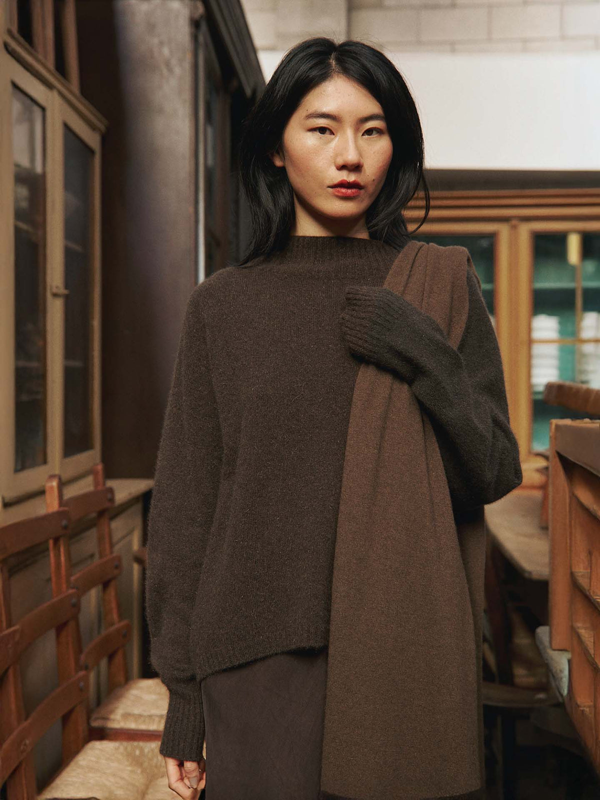 A woman with long dark hair wears the Fireside Knit – Truffle sweater in a relaxed fit and holds a matching scarf, standing in a room with wooden chairs and cabinets.