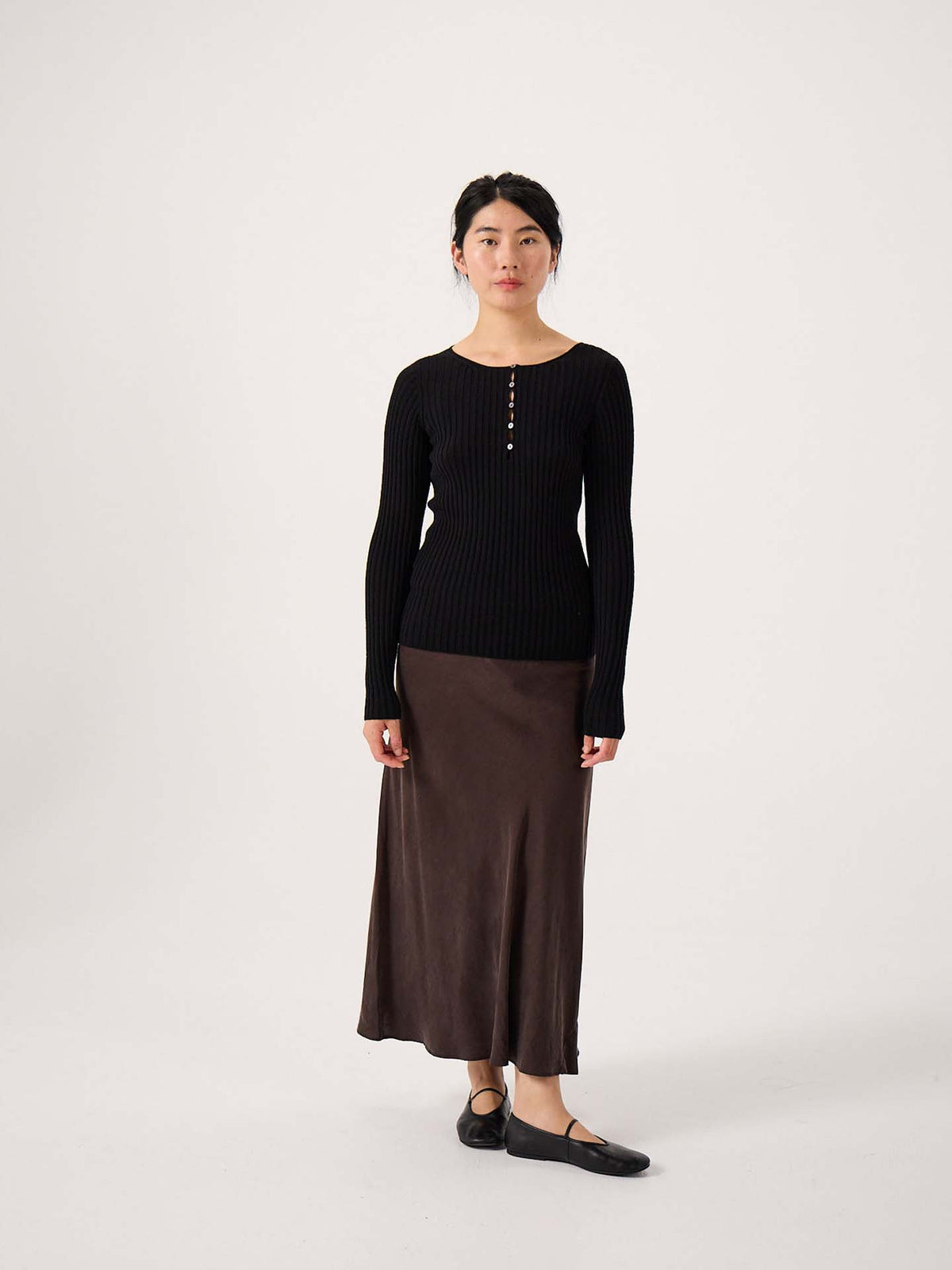 A woman stands against a plain background wearing the Form Top – Black, paired with a brown ankle-length skirt and black shoes.