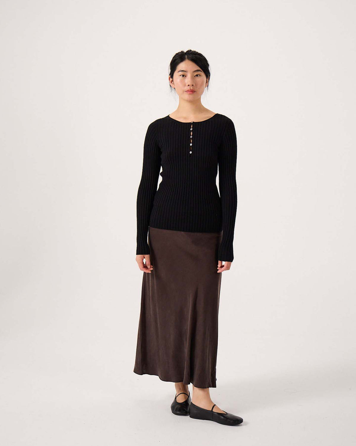 A woman stands against a plain background wearing the Form Top – Black, paired with a brown ankle-length skirt and black shoes.