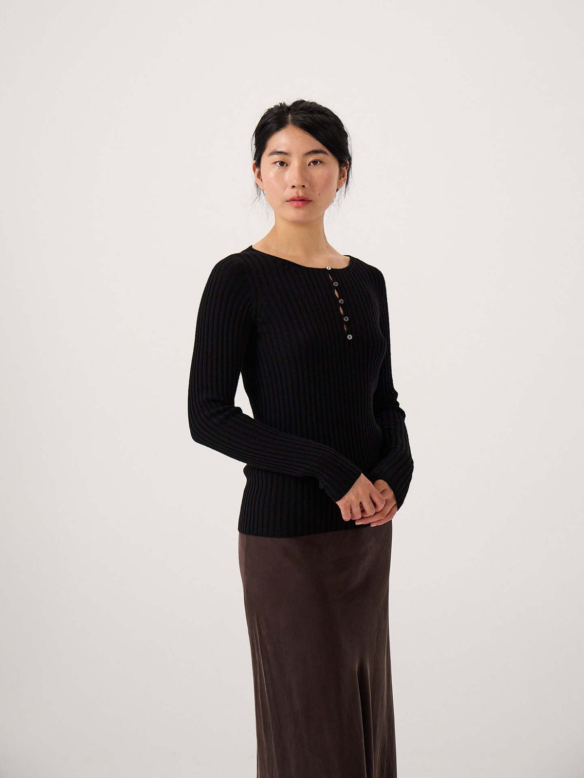 A woman stands against a plain background wearing the Form Top – Black, a ribbed long-sleeve top with buttons, paired with a brown skirt. She faces the camera with her hands clasped.