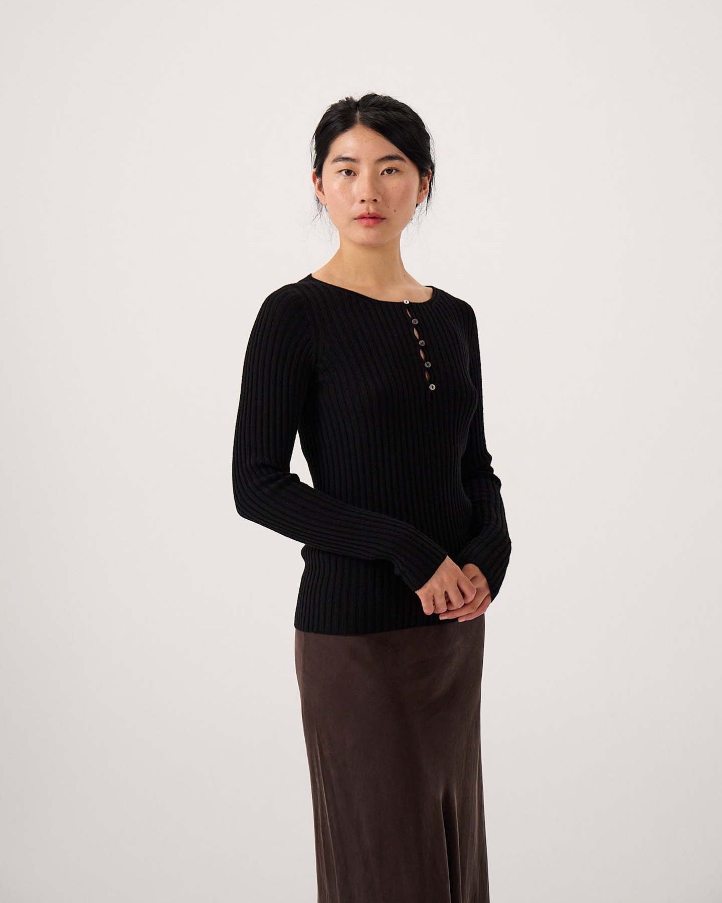 A woman stands against a plain background wearing the Form Top – Black, a ribbed long-sleeve top with buttons, paired with a brown skirt. She faces the camera with her hands clasped.