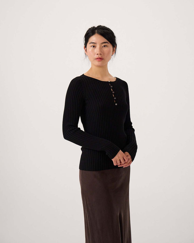 A woman stands against a plain background wearing the Form Top – Black, a ribbed long-sleeve top with buttons, paired with a brown skirt. She faces the camera with her hands clasped.