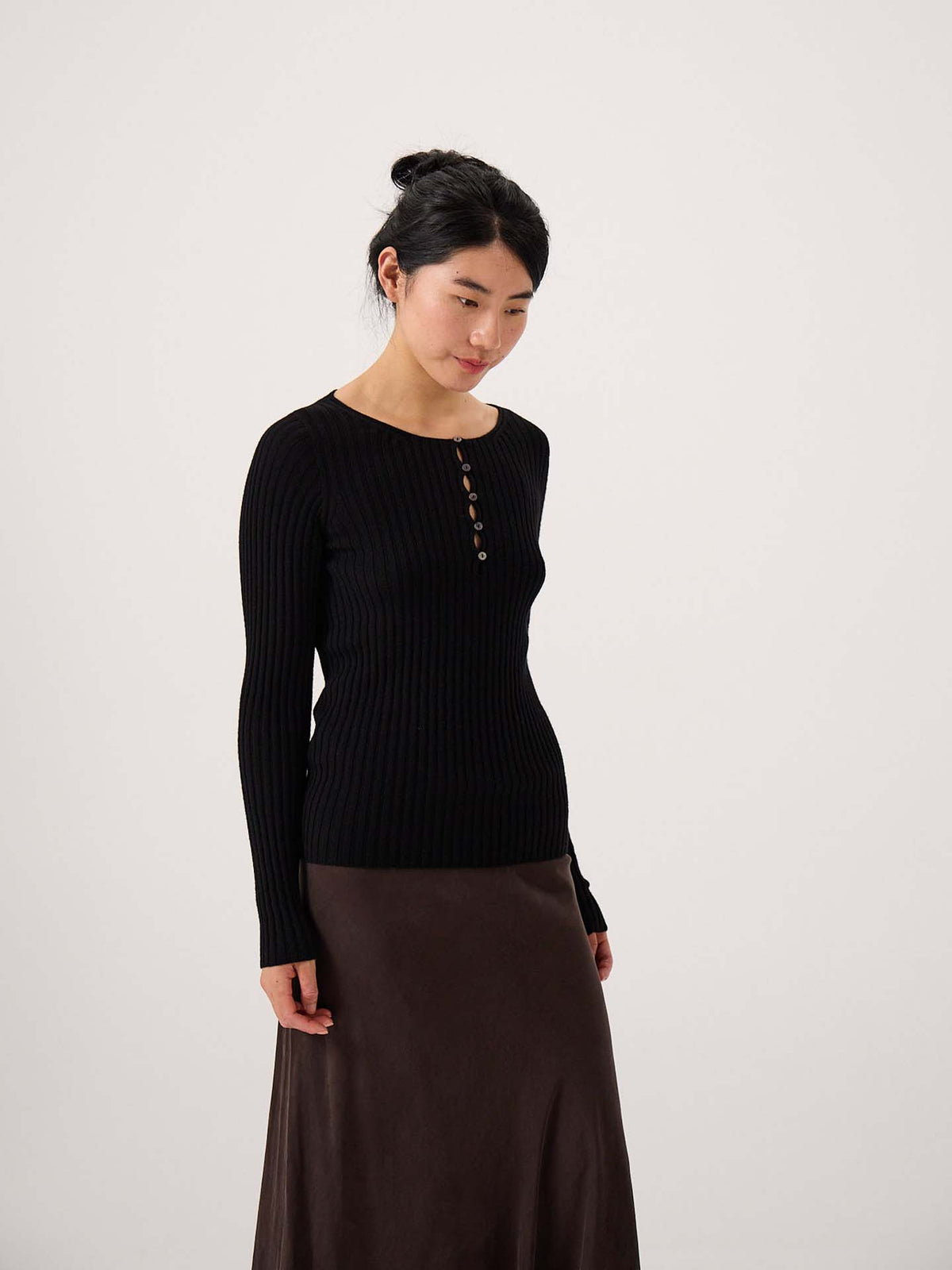 A woman stands against a plain background wearing the Form Top – Black, a long-sleeve ribbed top with buttons, paired with a dark brown skirt for a relaxed look.