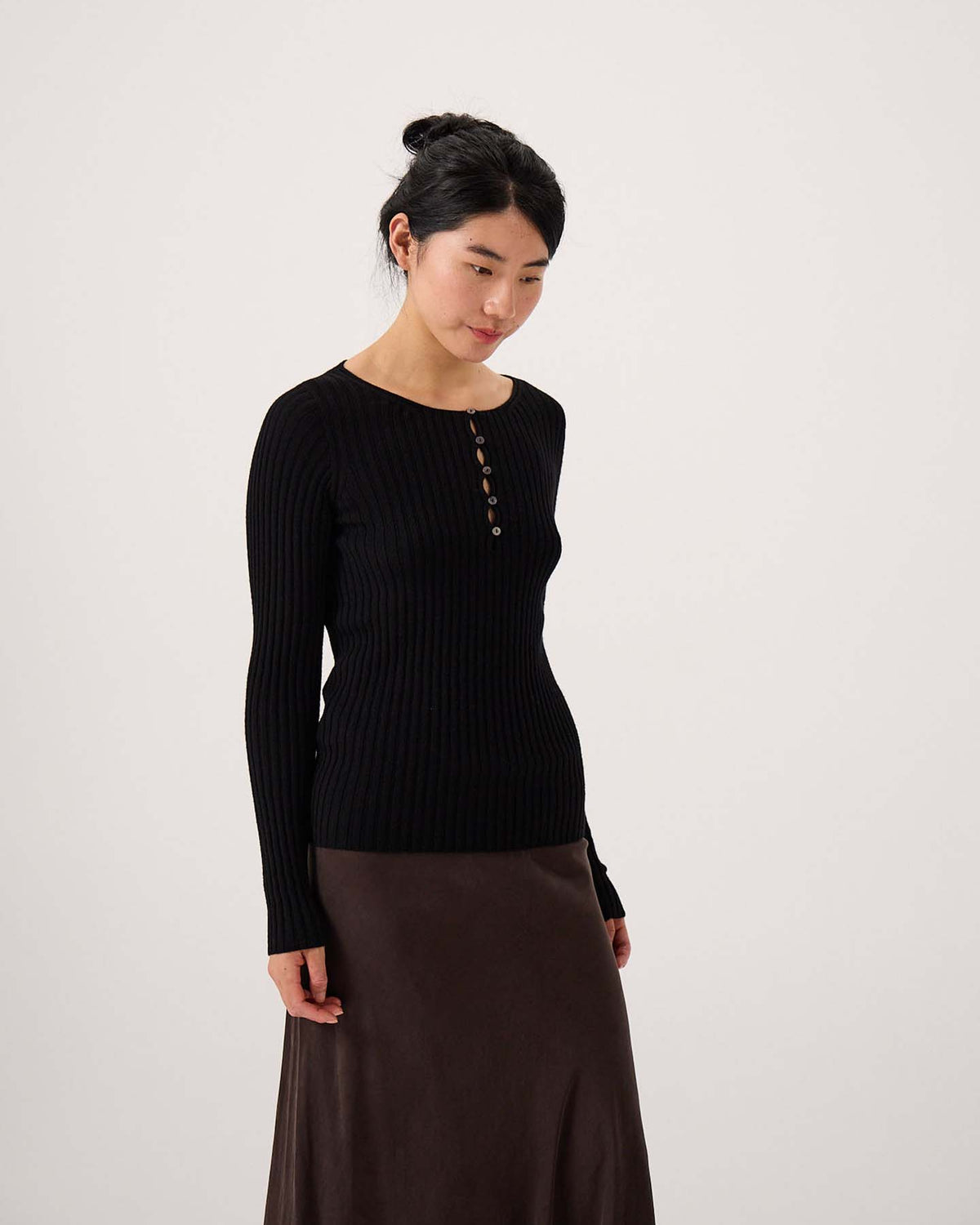 A woman stands against a plain background wearing the Form Top – Black, a long-sleeve ribbed top with buttons, paired with a dark brown skirt for a relaxed look.