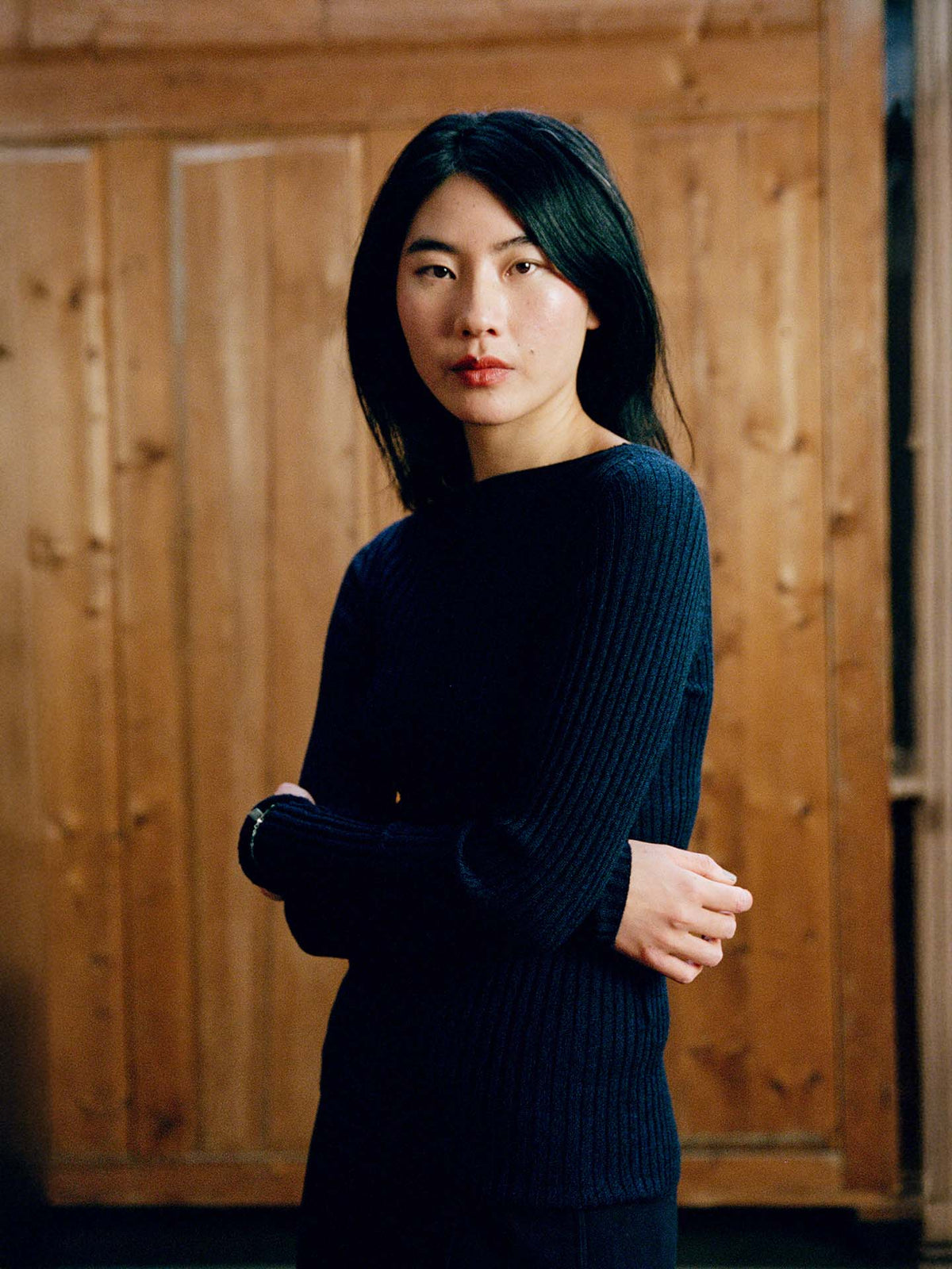A woman with long dark hair stands indoors before wooden panels, wearing the Form Top – Ink, a dark ribbed sweater, looking at the camera with her arms loosely crossed.