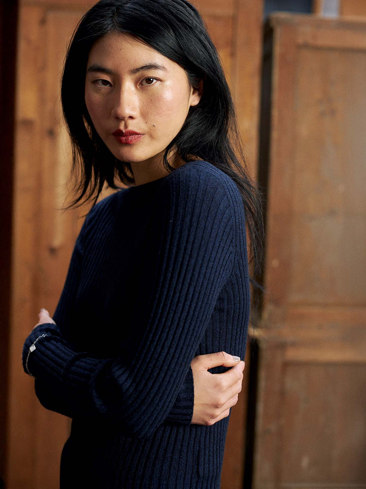 A person with long black hair wears the Form Top – Ink, a navy ribbed relaxed-fit sweater, standing indoors with arms crossed and looking at the camera.