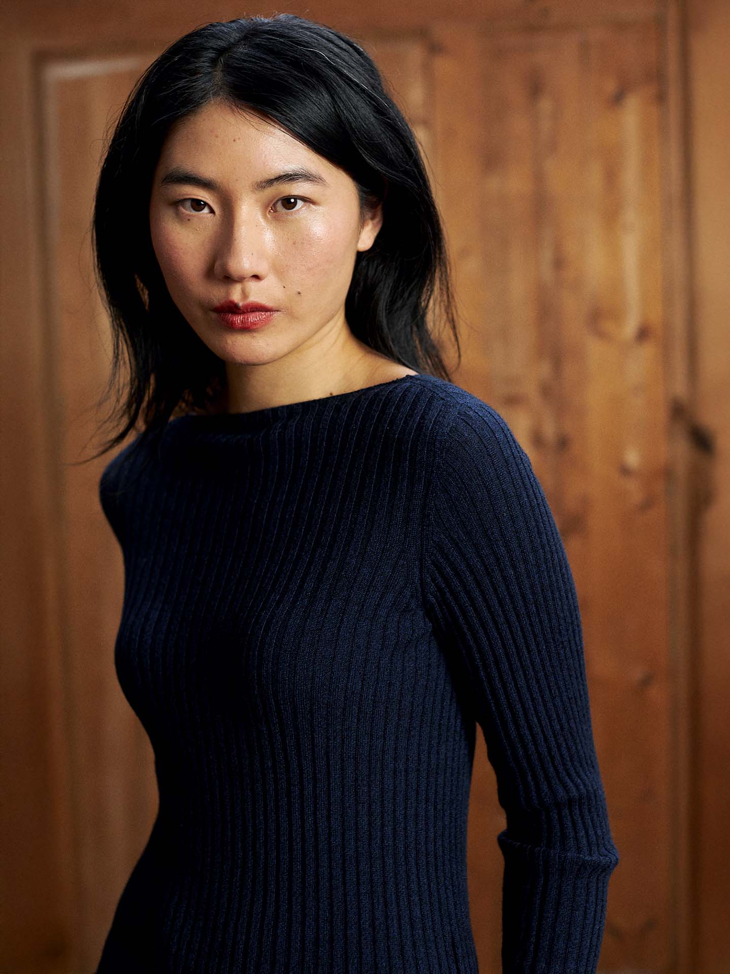 A woman with long black hair wears the Form Top – Ink, a ribbed navy blue sweater, standing in front of a wooden background and looking at the camera.