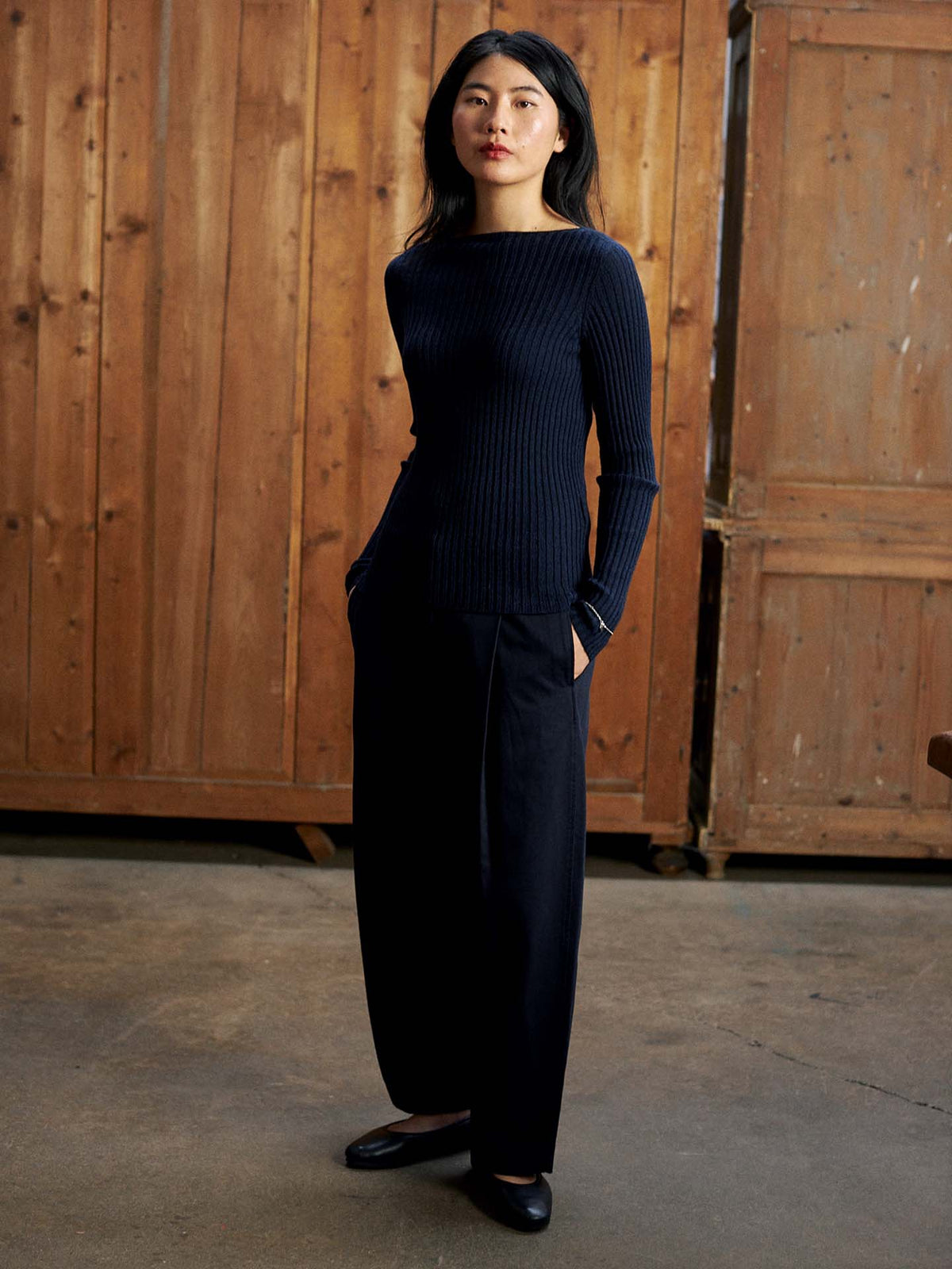 A woman stands indoors in front of a wooden wall and cabinet, wearing the Form Top – Ink, wide-leg black pants with a relaxed fit, and black shoes, her hands in her pockets.