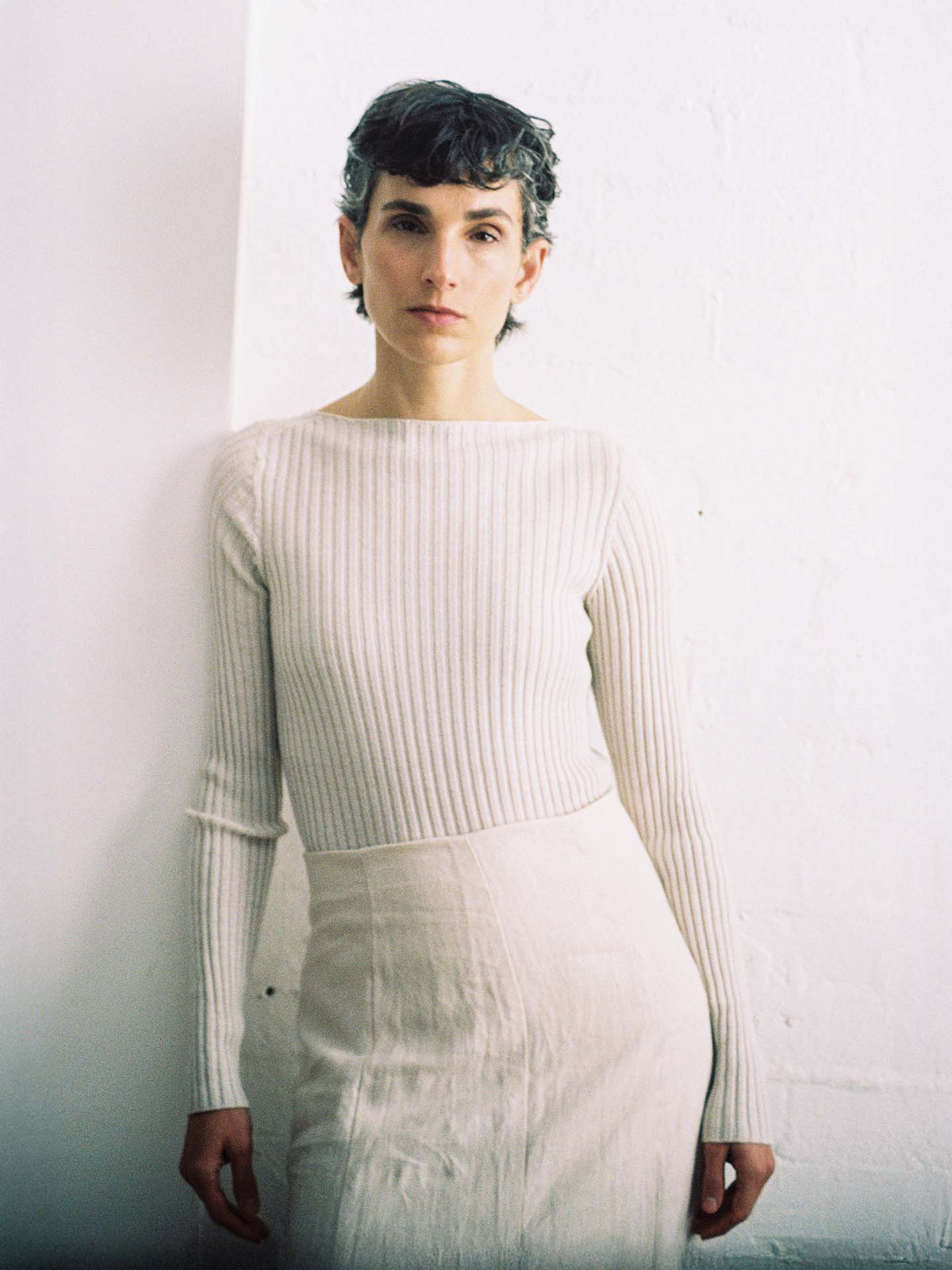 Person with short dark hair wears the Form Top – Ivory, a long-sleeved, ribbed white top, paired with a light skirt, standing against a plain white wall and looking at the camera.
