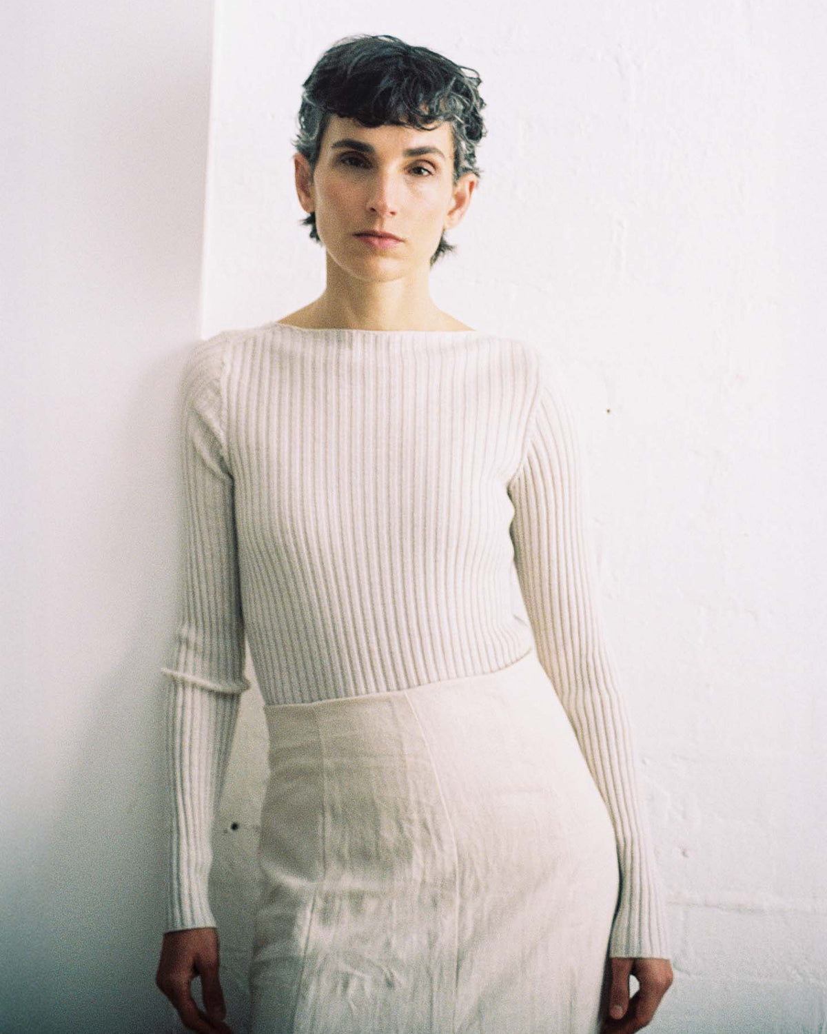 Person with short dark hair wears the Form Top – Ivory, a long-sleeved, ribbed white top, paired with a light skirt, standing against a plain white wall and looking at the camera.