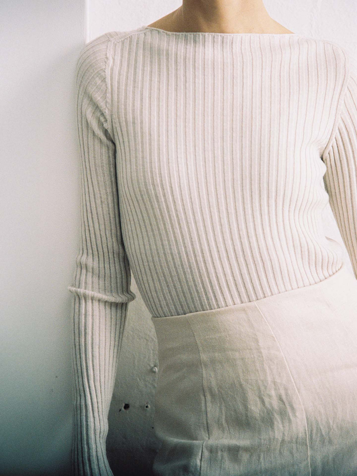 Person wearing the Form Top – Ivory, a long-sleeve, ribbed style top in a light shade, paired with a high-waisted, relaxed fit light skirt. Standing against a white wall with face not visible.