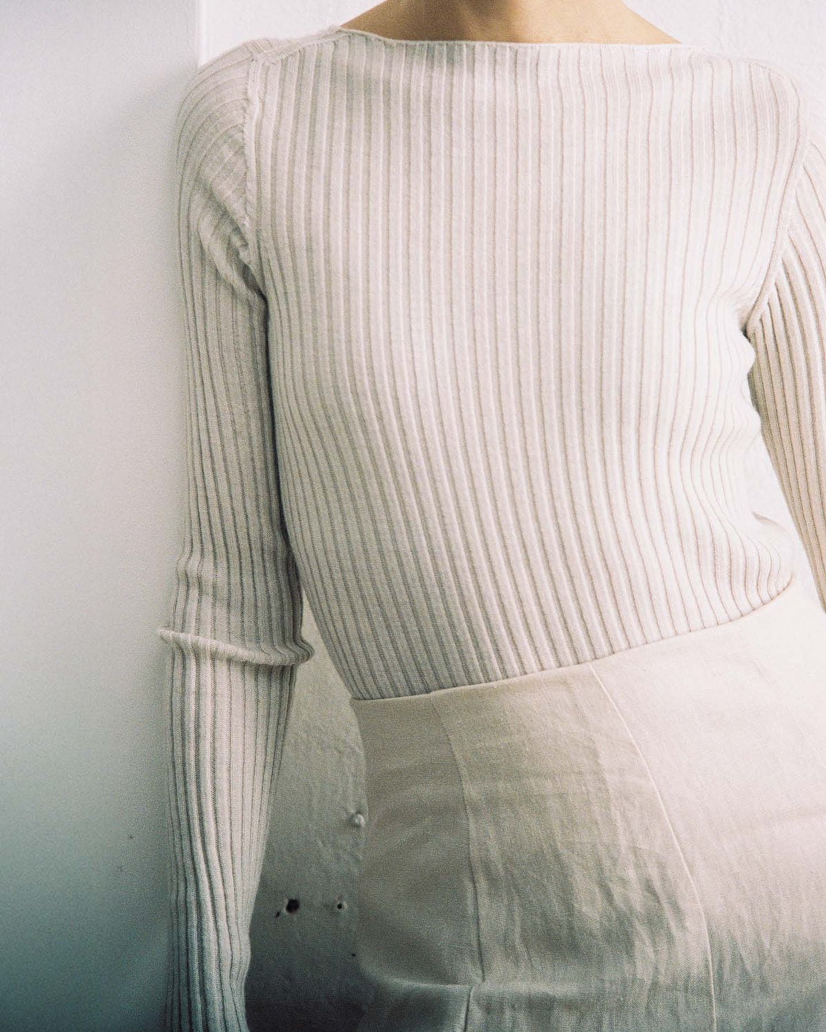 Person wearing the Form Top – Ivory, a long-sleeve, ribbed style top in a light shade, paired with a high-waisted, relaxed fit light skirt. Standing against a white wall with face not visible.