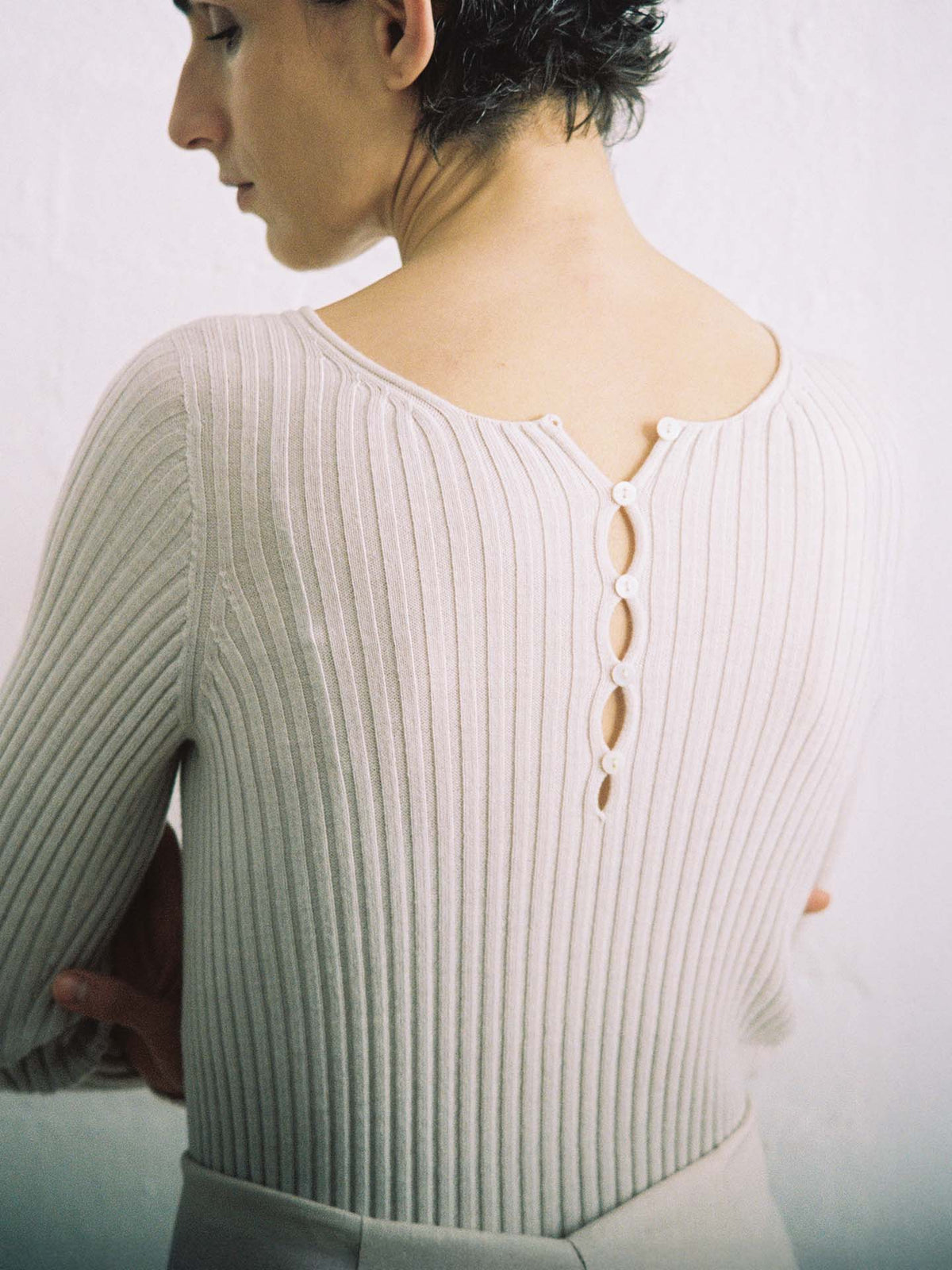 A person with short dark hair, seen from behind, wears the Form Top – Ivory: a ribbed, light-colored sweater with four back buttons. Its relaxed fit flatters crossed arms against a plain, light background.