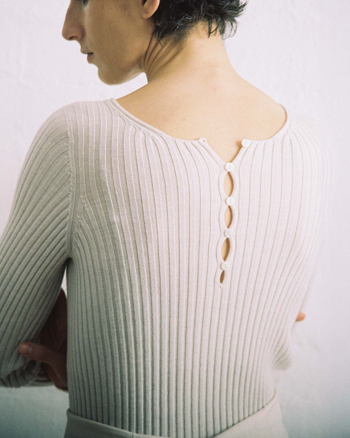 A person with short dark hair, seen from behind, wears the Form Top – Ivory: a ribbed, light-colored sweater with four back buttons. Its relaxed fit flatters crossed arms against a plain, light background.