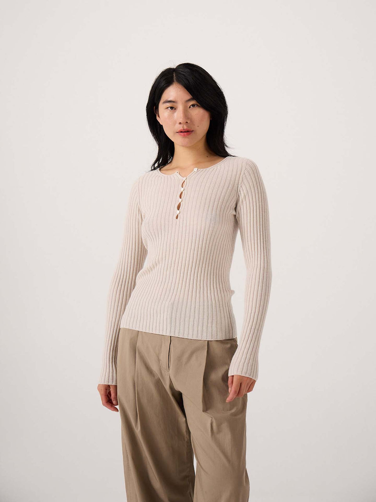 A petite person stands against a plain background, wearing the Form Top in Ivory, a stretch ribbed long-sleeve style with buttons, paired with loose tan pants.
