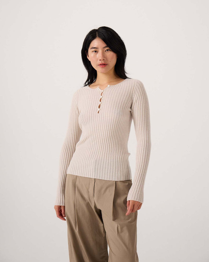A petite person stands against a plain background, wearing the Form Top in Ivory, a stretch ribbed long-sleeve style with buttons, paired with loose tan pants.