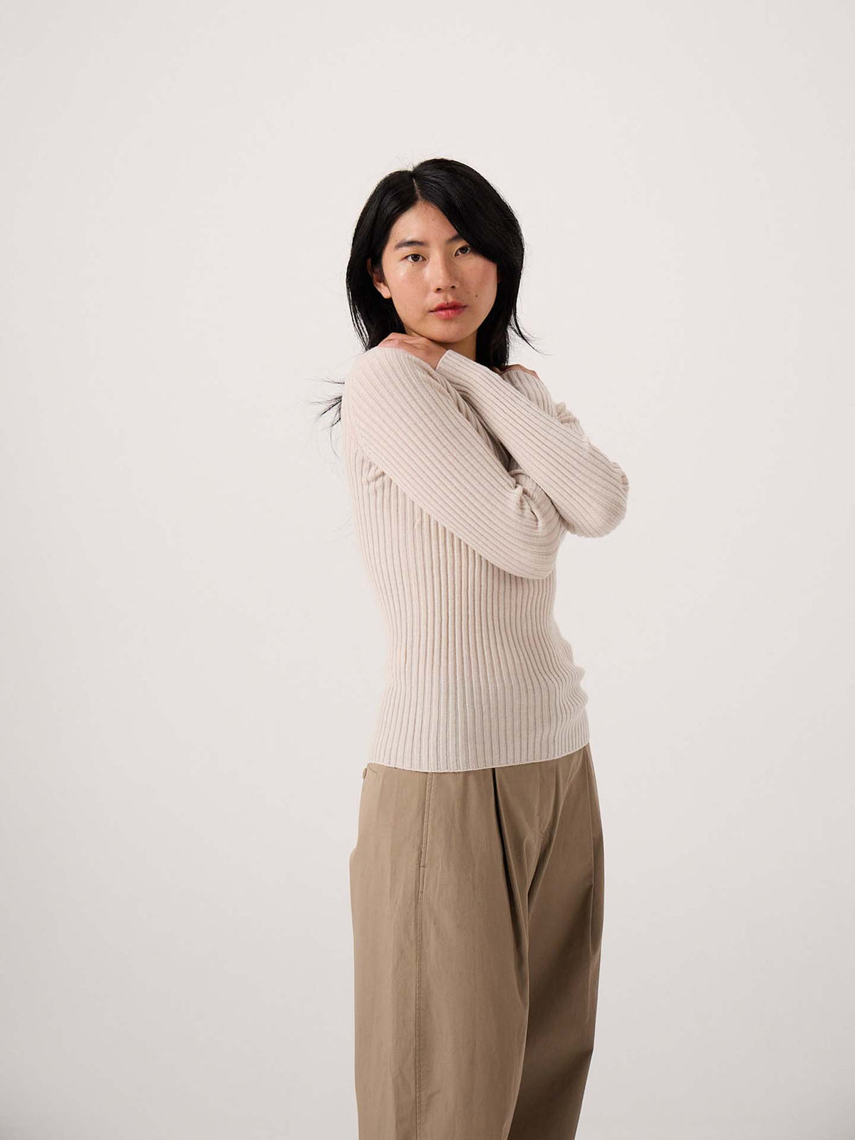 A petite woman with long black hair wears the Form Top – Ivory and loose tan pants, standing against a plain white background with her arms crossed.