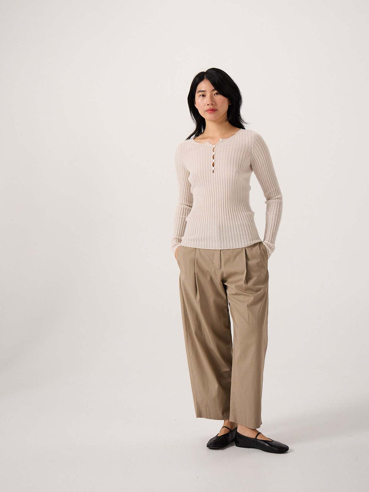 A petite woman stands against a plain background wearing the Form Top – Ivory with loose tan trousers and black flats, her hands in her pockets.